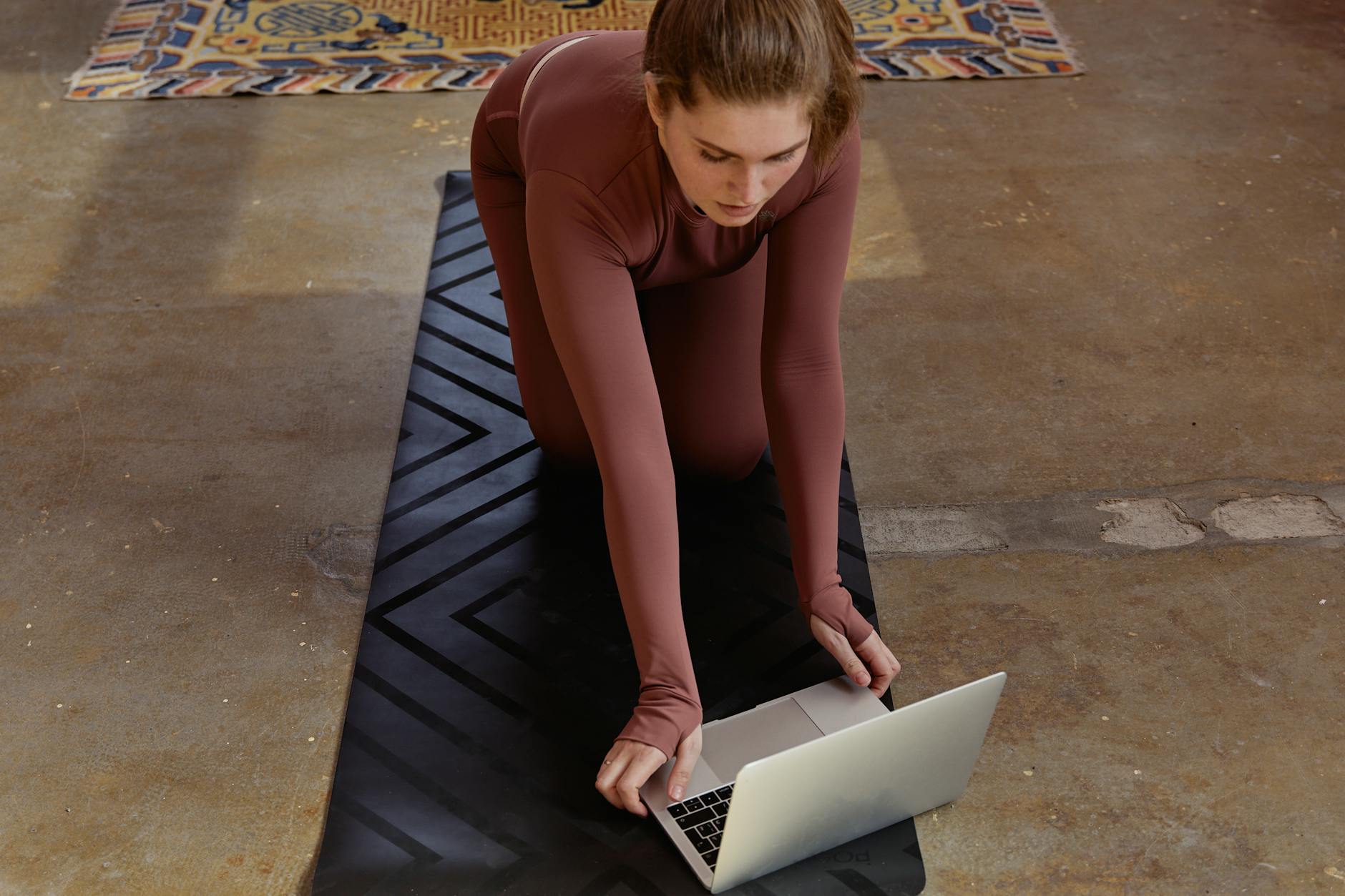 Woman in yoga attire kneeling on mat using laptop indoors, blending fitness with technology.
