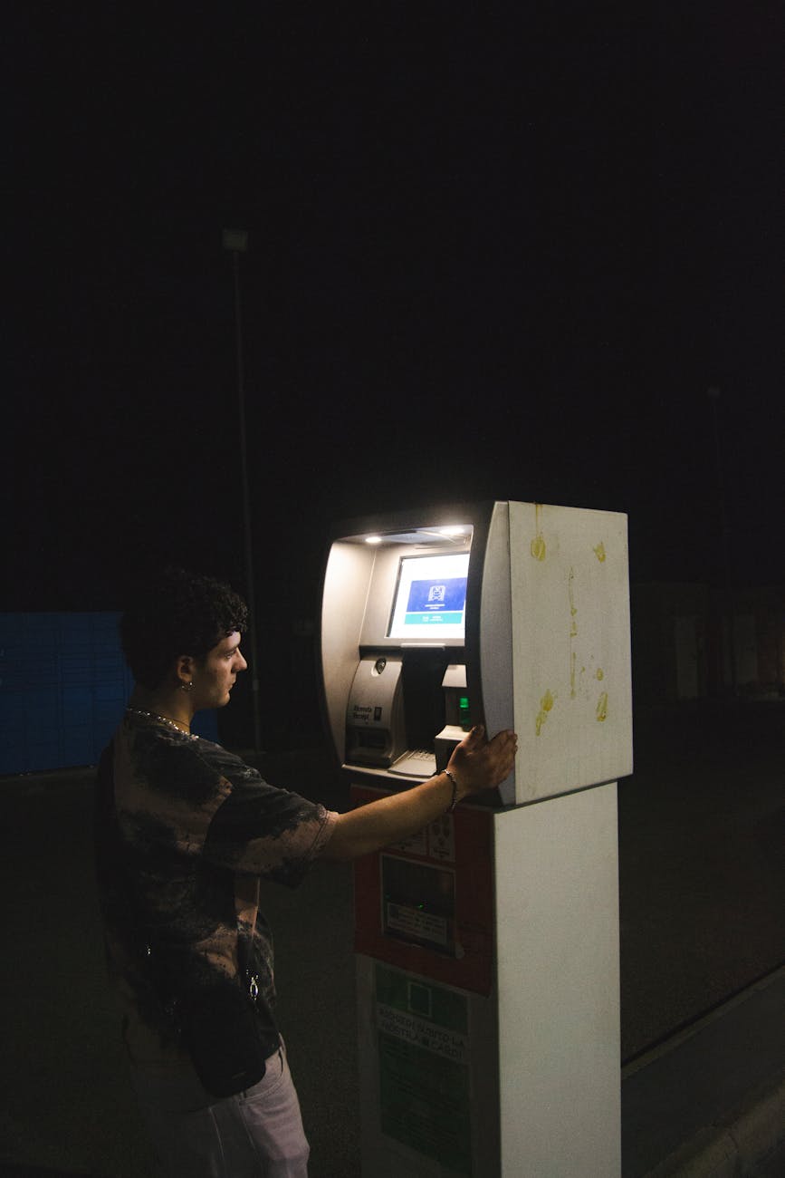 A man uses an ATM on a dimly lit street at night, highlighting finance technology.
